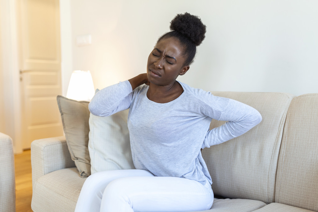 Young Black woman suffering from backache at home. Portrait of a young girl sitting on the couch at home with a headache and back pain. Beautiful woman Having Spinal Or Kidney Pain