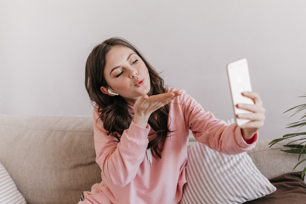 Woman in sweatshirt and wireless headphones blows kiss and takes selfie. Portrait of brunette curly teen girl in pink outfit talking by video call at home