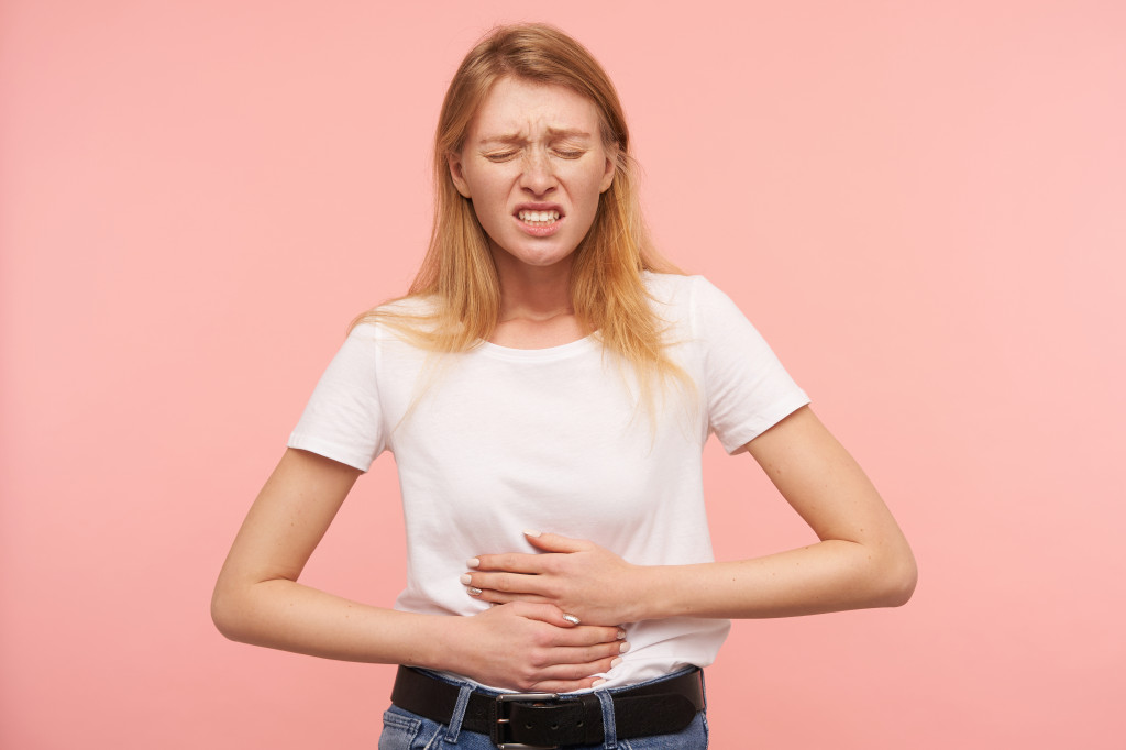 Indoor photo of young pretty redhead lady keeping her eyes closed and frowning face while having stomachache, standing over pink background in casual clothes