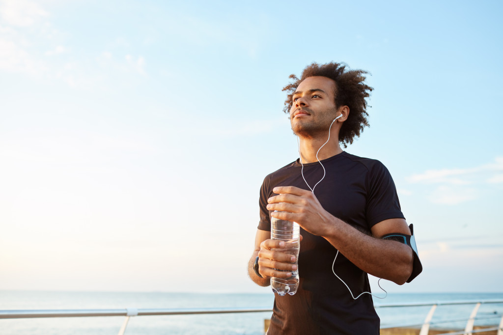 Man athlete drinking water out of plastic bottle after hard running workout. Dark-skinned male sportsman looking at the sky while running, enjoying view