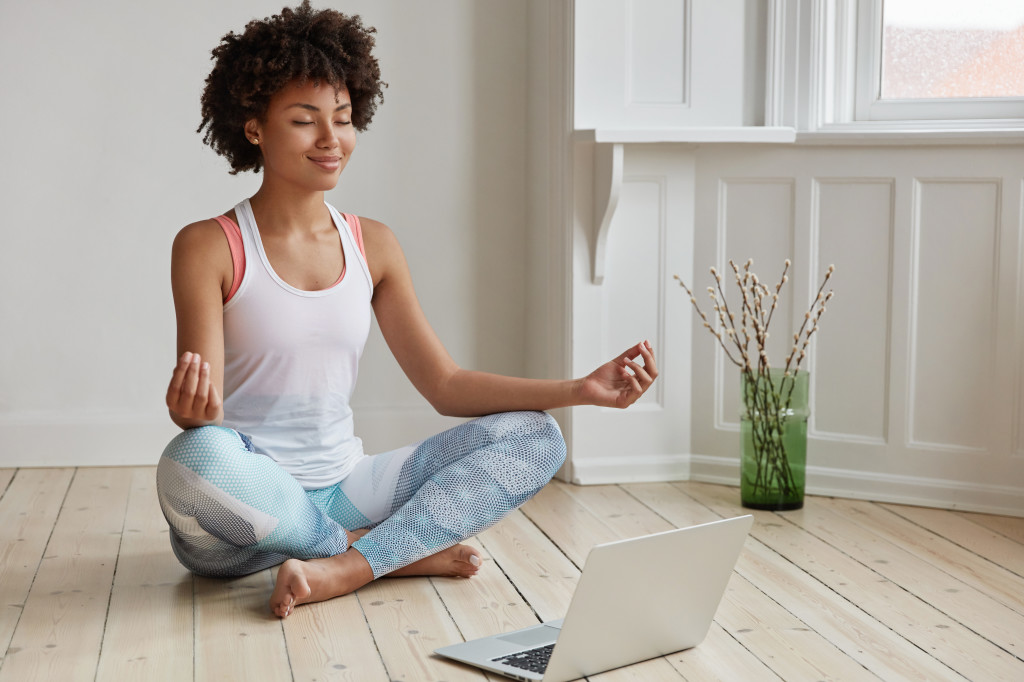 People, ethnicity, yoga and meditation concept. Beautiful pleased delighted dark skinned relaxed student sits crossed legs on floor near opened laptop, tries to have break after work on diploma paper