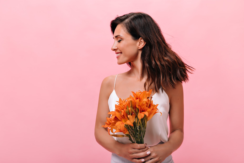 Adorable woman in top laughs and poses with bunch of flowers on pink background