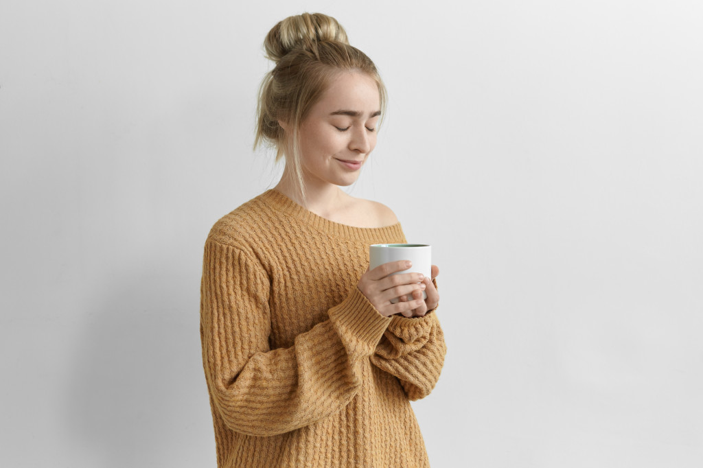 Isolated image of gorgeous charming young female with hair knot holding large mug enjoying freshly made cappuccino from large mug, wearing cozy oversize pullover, keeping eyes closed and smiling