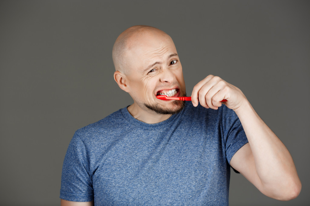 Portrait of funny handsome middle-aged man in grey shirt brushing teeth over dark background.