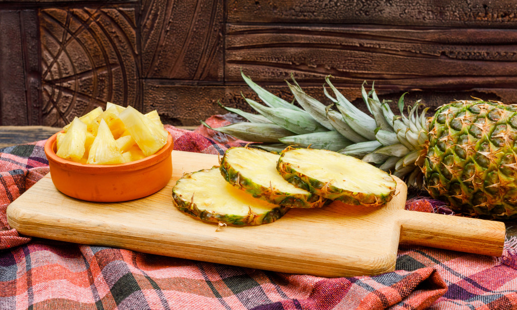 Whole and sliced pineapple in a cutting board and clay bowl on a stone tile and picnic cloth background. side view.