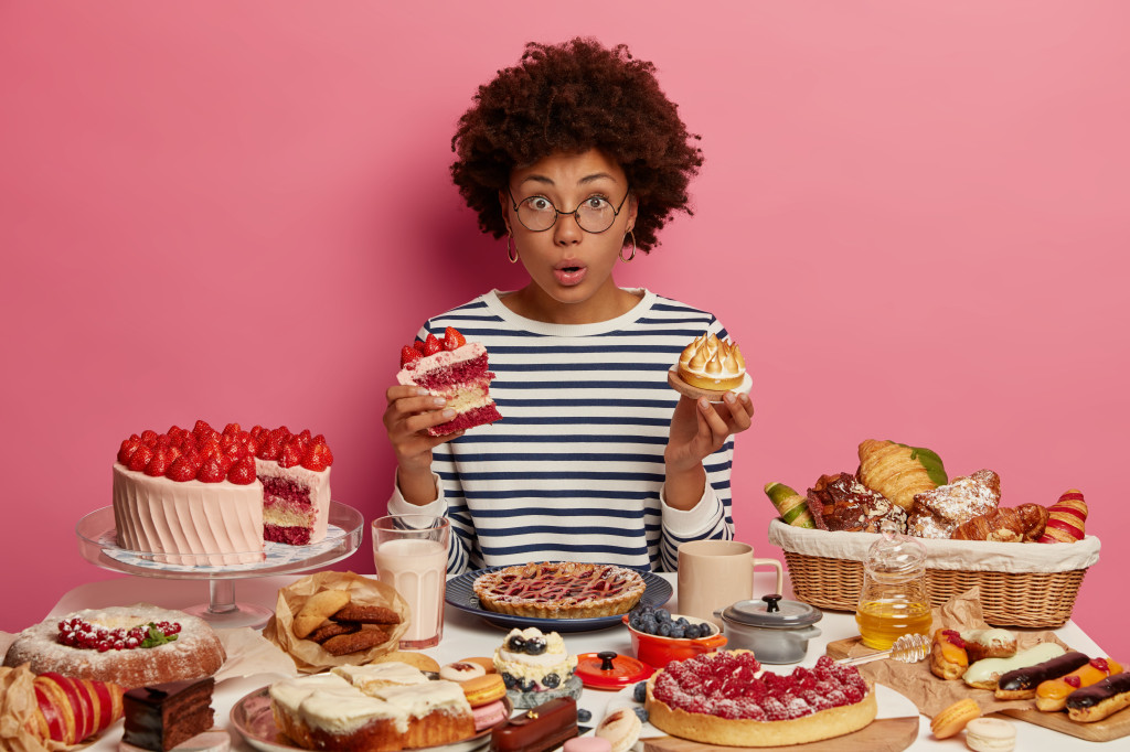 Emotional surprised dark skinned woman eats cake and cupcake, surrounded by tasty homemade desserts, has unhealthy nutrition, cannot believe something, isolated on pink wall. Junk food, calories