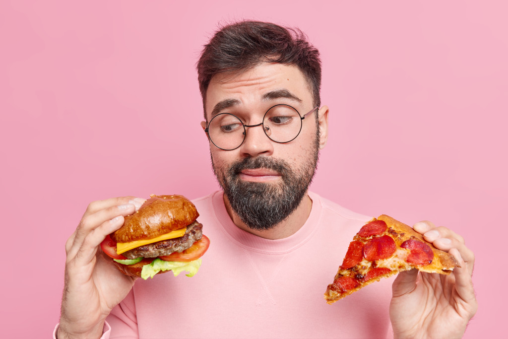 Handsome bearded adult European man feels hesitant whether to eat hamburger or pizza prefers eating junk food wears round spectacles and jumper isolated over pink background. Cheat meal concept