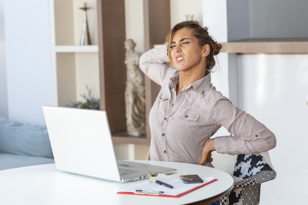 Portrait of young stressed woman sitting at home office desk in front of laptop, touching aching back with pained expression, suffering from backache after working on laptop