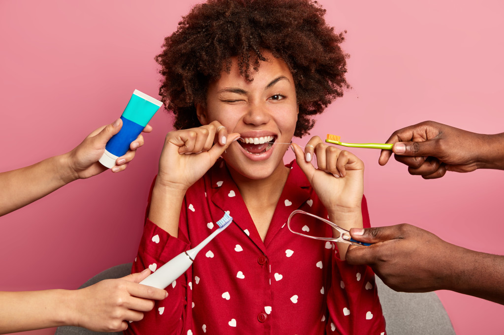 Happy curly young woman brushes teeth with tooth floss, cares about oral hygiene, surrounded with toothpaste, electric toothbrush and tongue cleaner, wears red pyjama, isolated on pink wall.